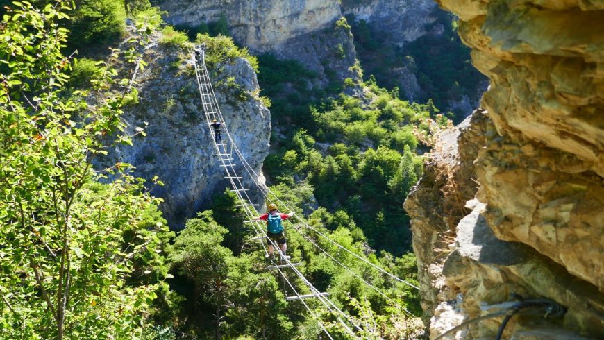 Eine schmale Hängebrücke überquert eine felsige Schlucht, umgeben von üppiger Vegetation und dramatischen Klippen.