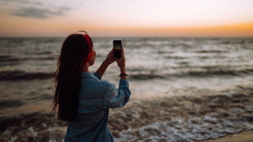 Junge Frau fotografiert das Meer bei Sonnenuntergang mit ihrem Smartphone, um am Ecobnb Regenerate-Wettbewerb teilzunehmen.