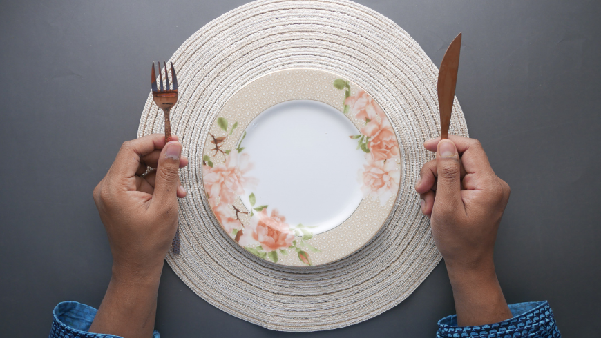 Person, die an einem Tisch sitzt und eine Gabel und ein Messer vor einem leeren Teller hält, was Hunger und Appetitkontrolle symbolisiert.