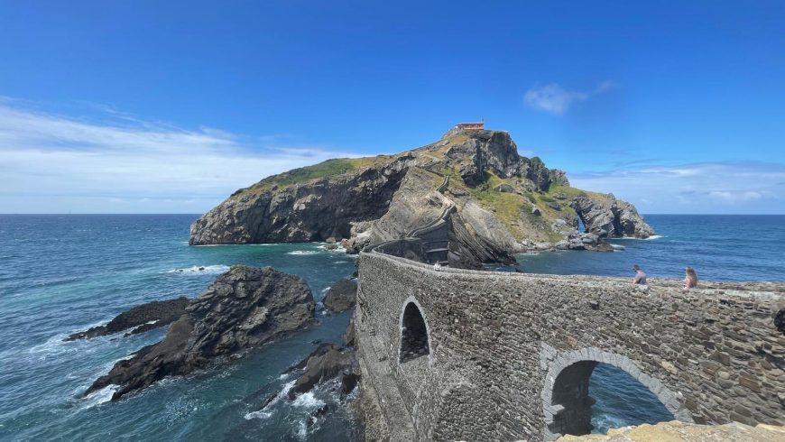 die Eremitage von San Juan de Gaztelugatxe mit der Steinbrücke im Vordergrund und zwei Menschen, die sie überqueren